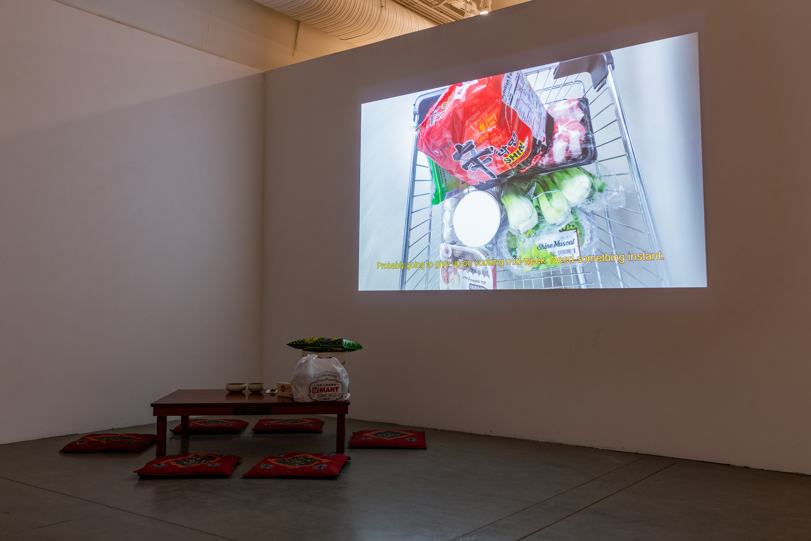 A dark white room with a low table on the left, with 6 seat cushions. On the table are groceries and two bowls. On the right is a projection of a video.